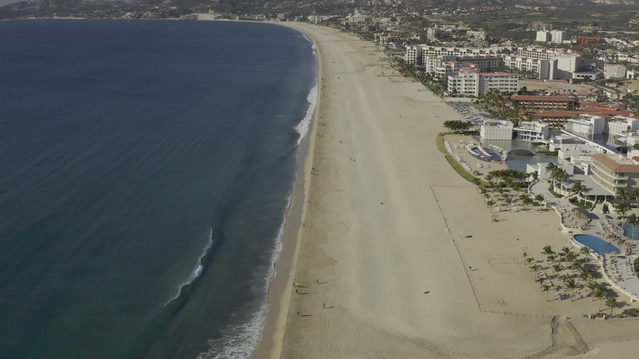 Aerial view along the Baja California Sur shoreline, with the ocean curving beside a broad sand beach and clustered beachfront buildings visible to the right with no visible people.
