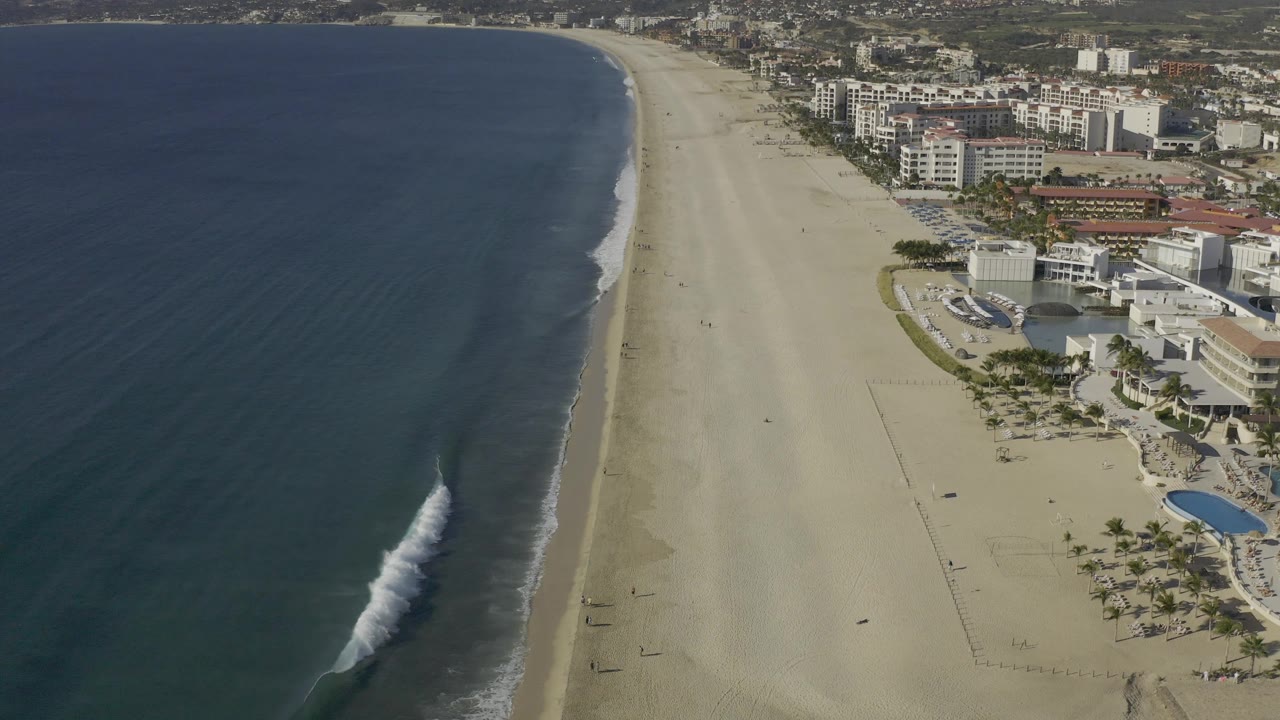 High drone view over Baja California Sur, Mexico, showing a brighter white wave breaking near the left edge of the sand while hotels or residential buildings line the right side, with no visible people.