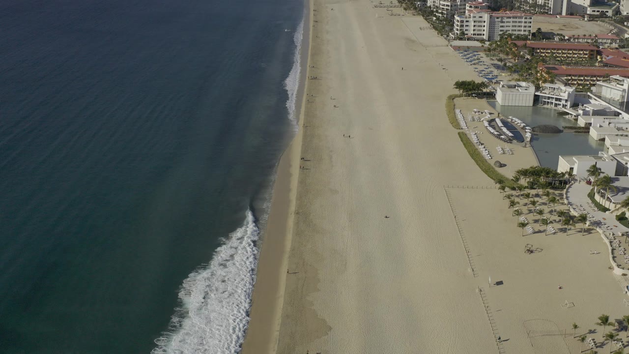 High aerial frame over Baja California Sur, Mexico showing dark ocean at left, a pale beach through the center, surf on the shore, and beachfront buildings at right, with no visible people.