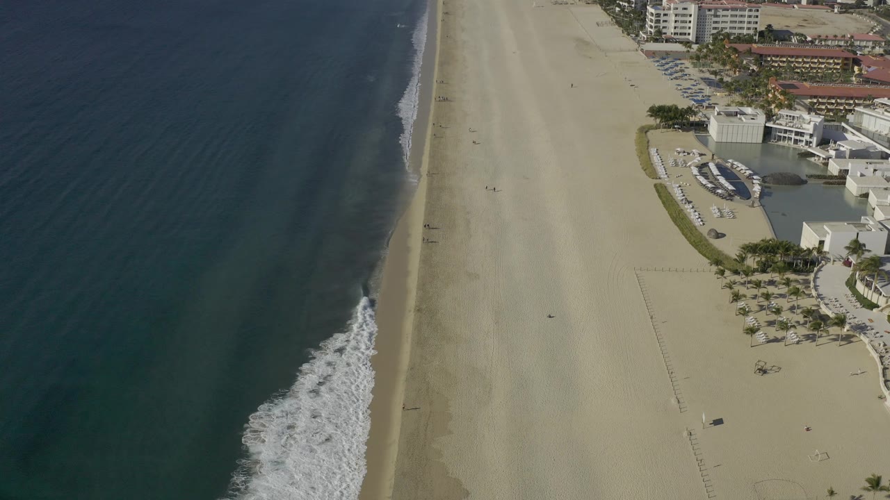 High drone-style frame over Baja California Sur, Mexico showing blue water on the left, a narrow white wave edge, broad sand, and a row of beachfront structures at the right edge, with no visible people.