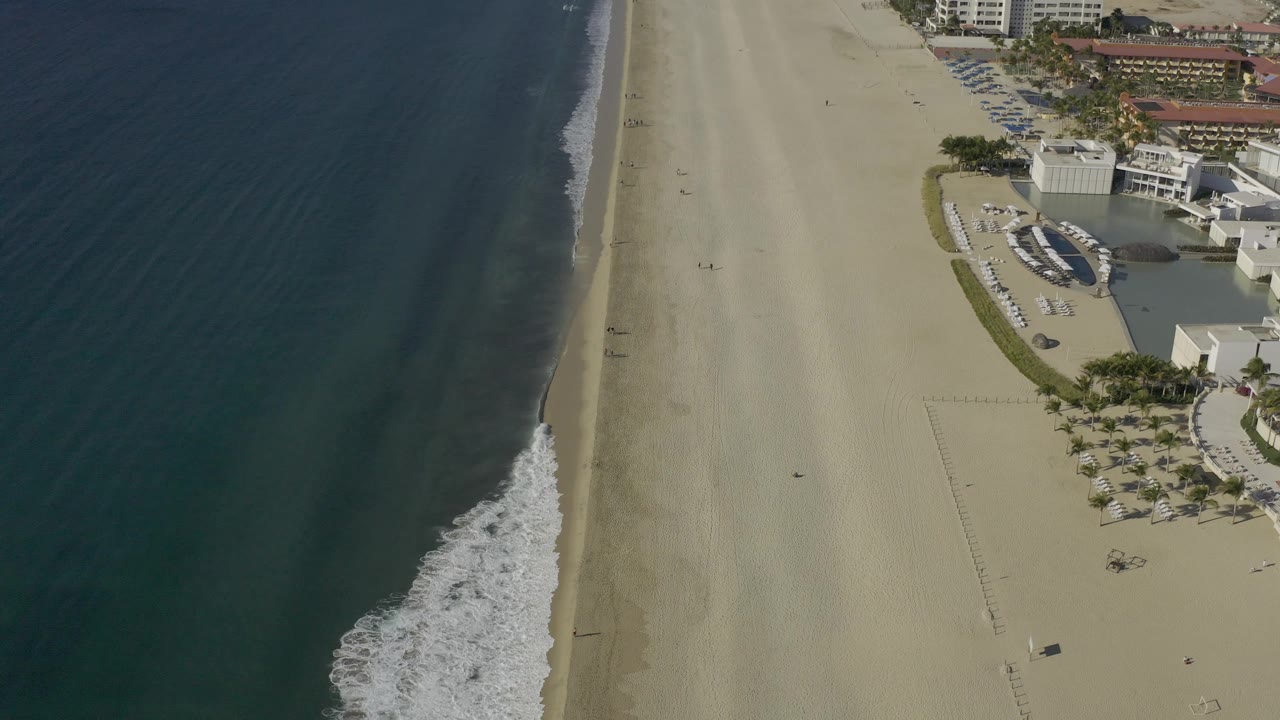 Aerial beach frame with ocean on the left and resort-like buildings on the right.