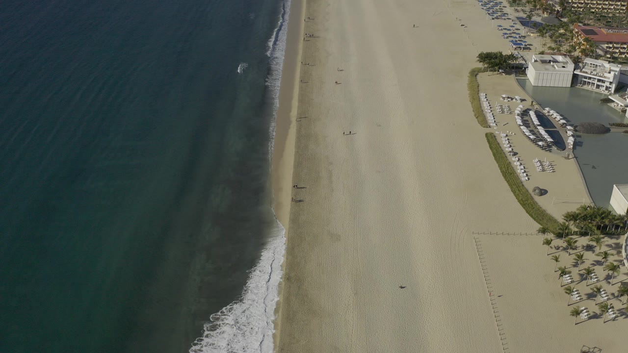 Aerial frame in Baja California Sur, Mexico showing dark ocean beside a wide pale sand beach and a beachfront amenity area at the right edge, with no visible people.