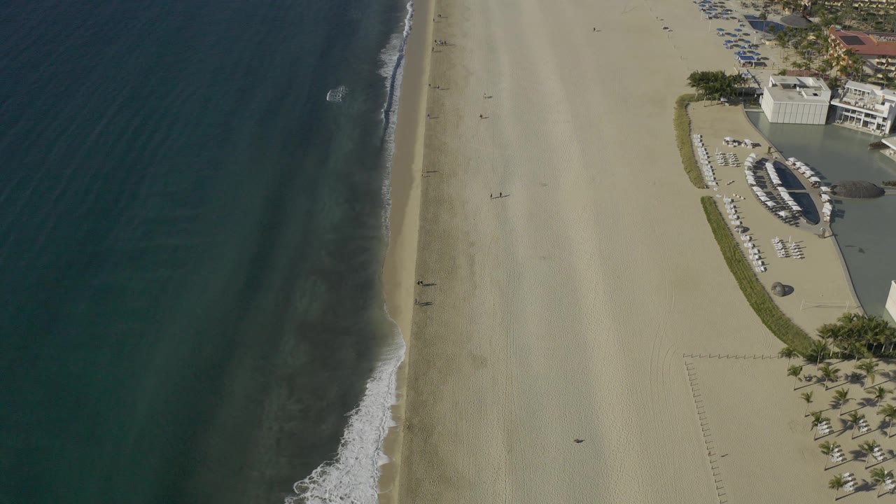 Aerial Baja California Sur coastline frame with darker bands in the water, a narrow breaking wave, open sand, and rows of loungers beside buildings on the right, with no visible people.