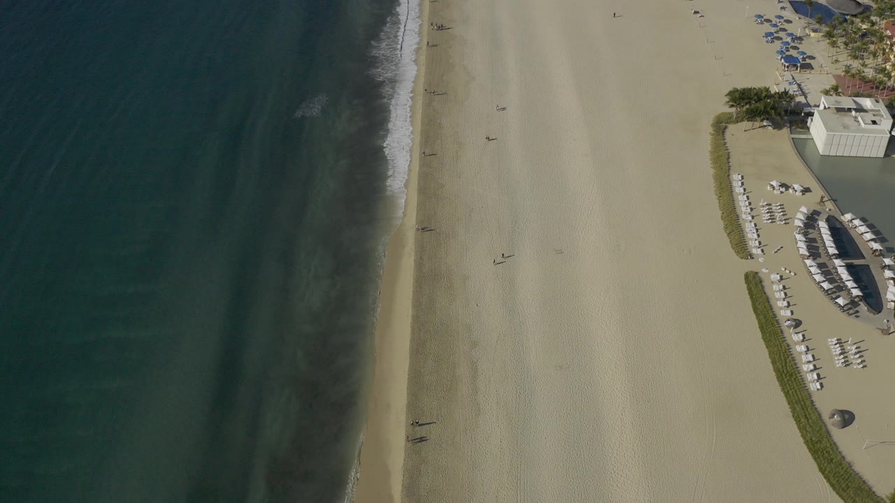 Overhead view of Baja California Sur beach with dark teal water on the left, pale sand in the center, and rows of beach loungers near a building at the right edge, with no recognizable people visible.