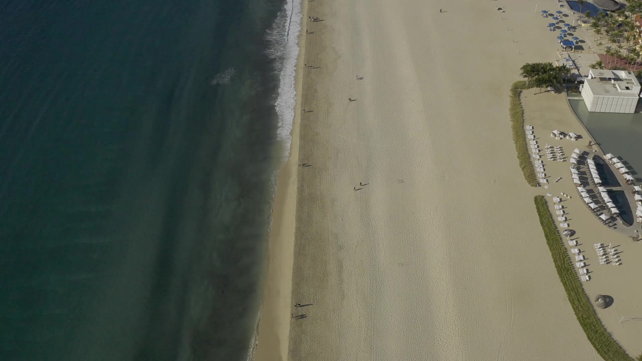 Aerial shoreline with dark water, narrow foam, open sand, and beach chairs near the right edge.