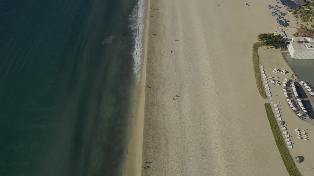 Top-down Baja California Sur beach view with darker water bands, a thin surf edge, smooth pale sand, and blue umbrellas gathered near the upper right, with no recognizable people visible.