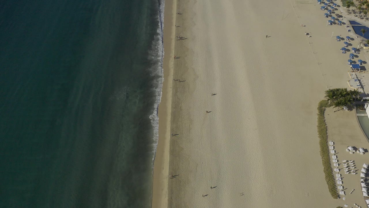 Overhead drone frame of Baja California Sur shoreline with dark green ocean at left, a narrow surf line, pale low tide sand, and blue beach umbrellas near the upper right.