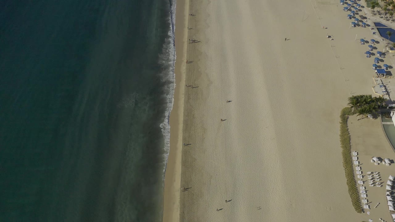Aerial beach frame with dark water, low tide sand, and umbrella rows.