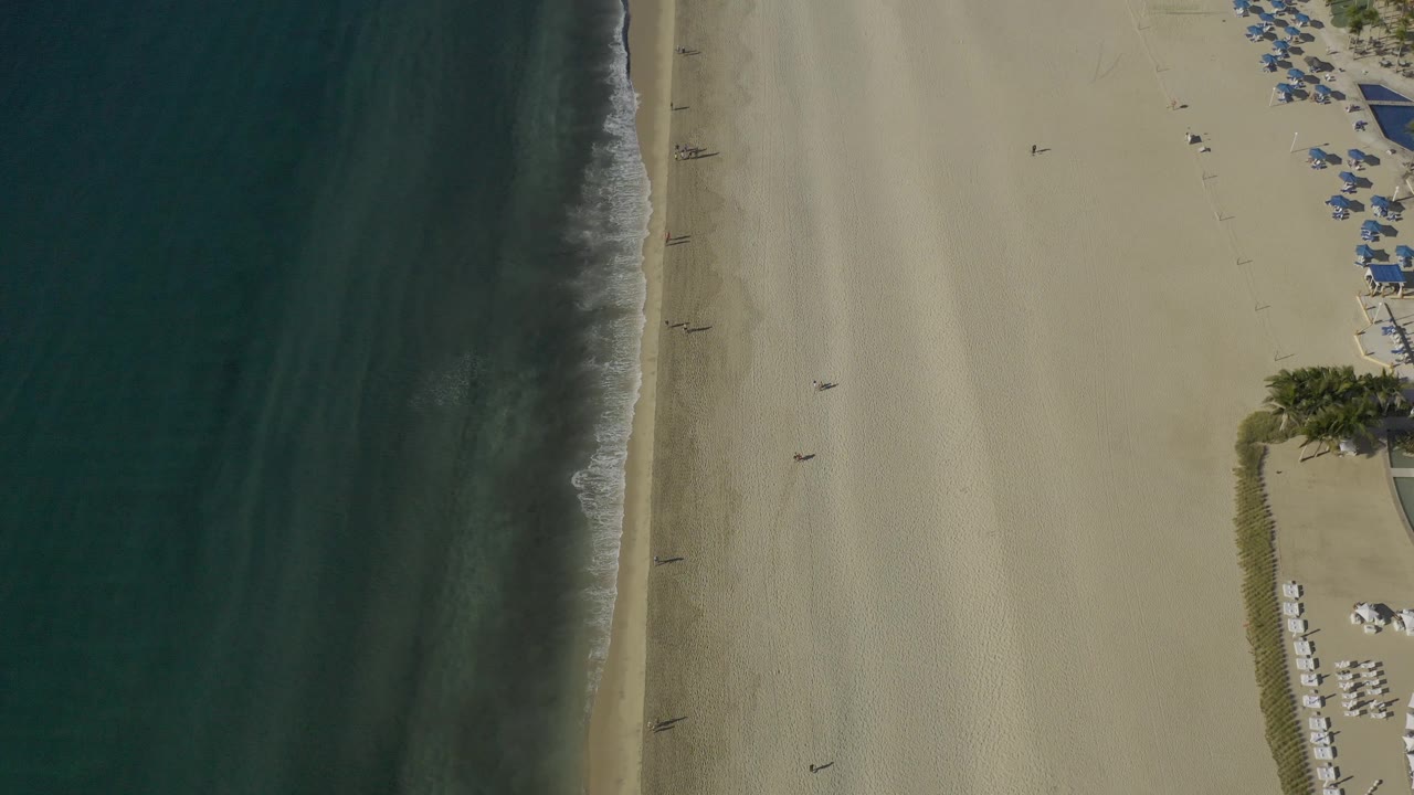 Aerial frame over the East Cape coastline with dark water filling the left side, pale sand dominating the center, and umbrella rows partly visible at the upper right edge.
