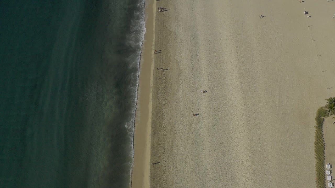 Top-down aerial view in Baja California Sur, Mexico, with dark turquoise water at left, a thin surf line, broad sand, tiny people, and a small beach setup near vegetation at lower right.