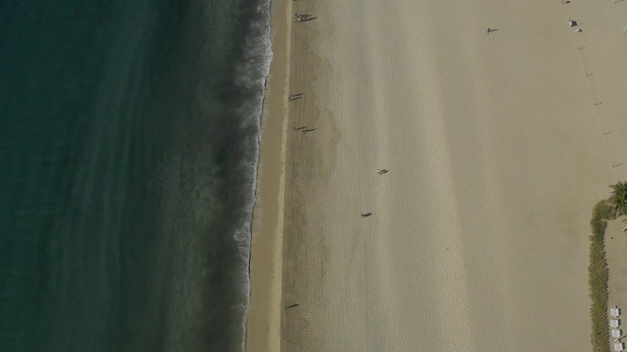 Overhead Baja California Sur beach frame with the ocean occupying the left side, pale sand on the right, scattered tiny people, and the beach setup still visible near the lower right edge.