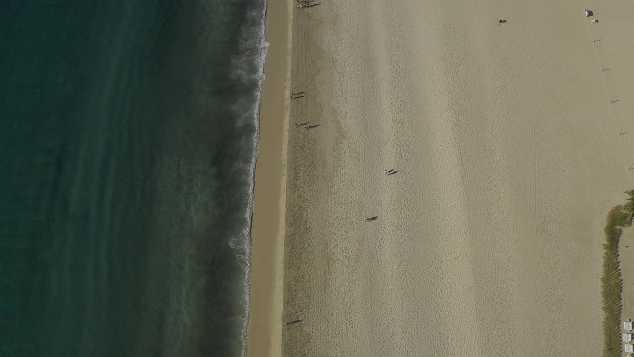 Top-down aerial frame in Baja California Sur, Mexico, showing textured turquoise water, a narrow breaking wave, wide sand, several small figures, and vegetation with beach items along the lower right edge.