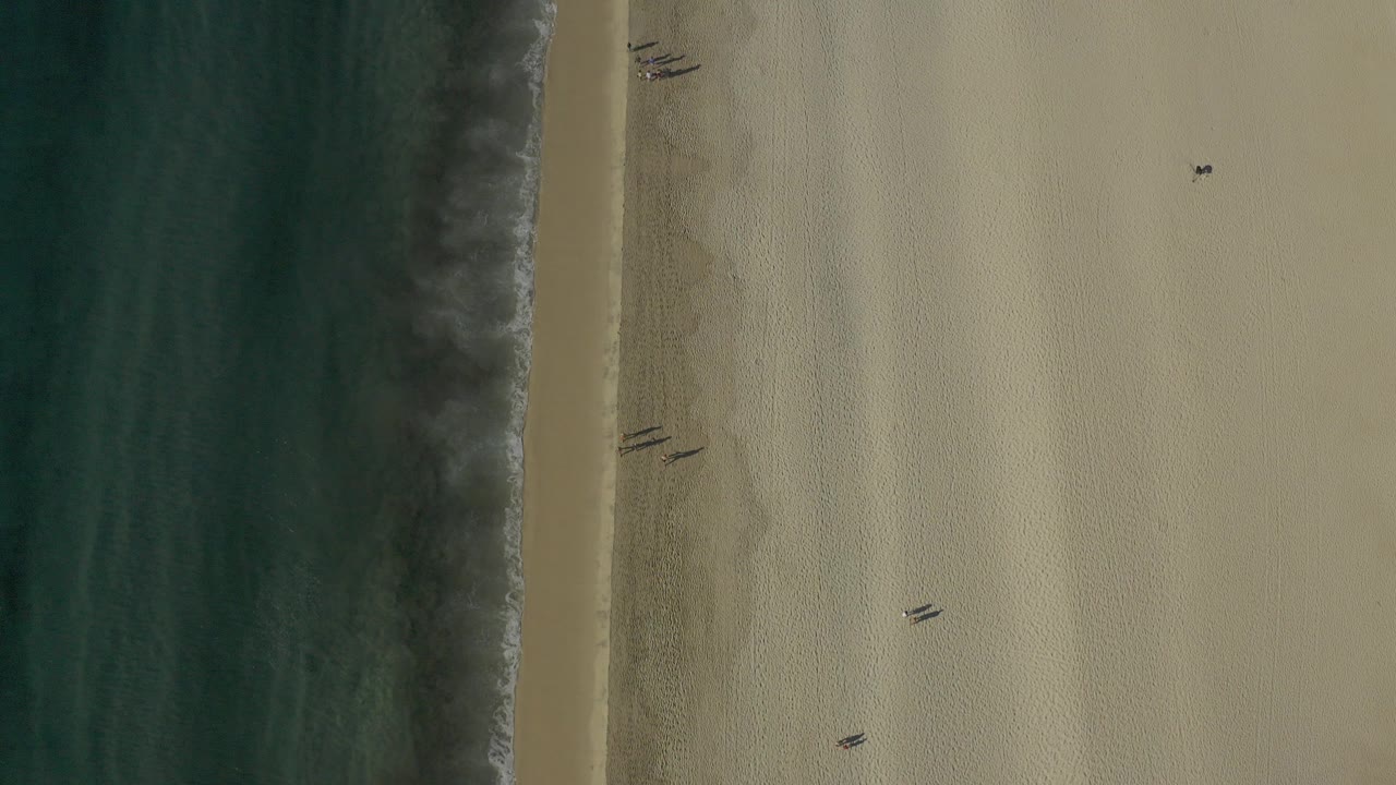 Overhead view of a Baja California Sur shoreline with dark turquoise water, a thin surf line, pale sand, scattered beach marks, and no visible people.