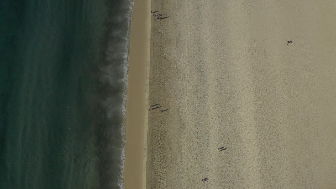 Top-down Baja California Sur beach frame showing the surf edge slightly farther into view, with broad pale sand, small dark marks, and no visible people.