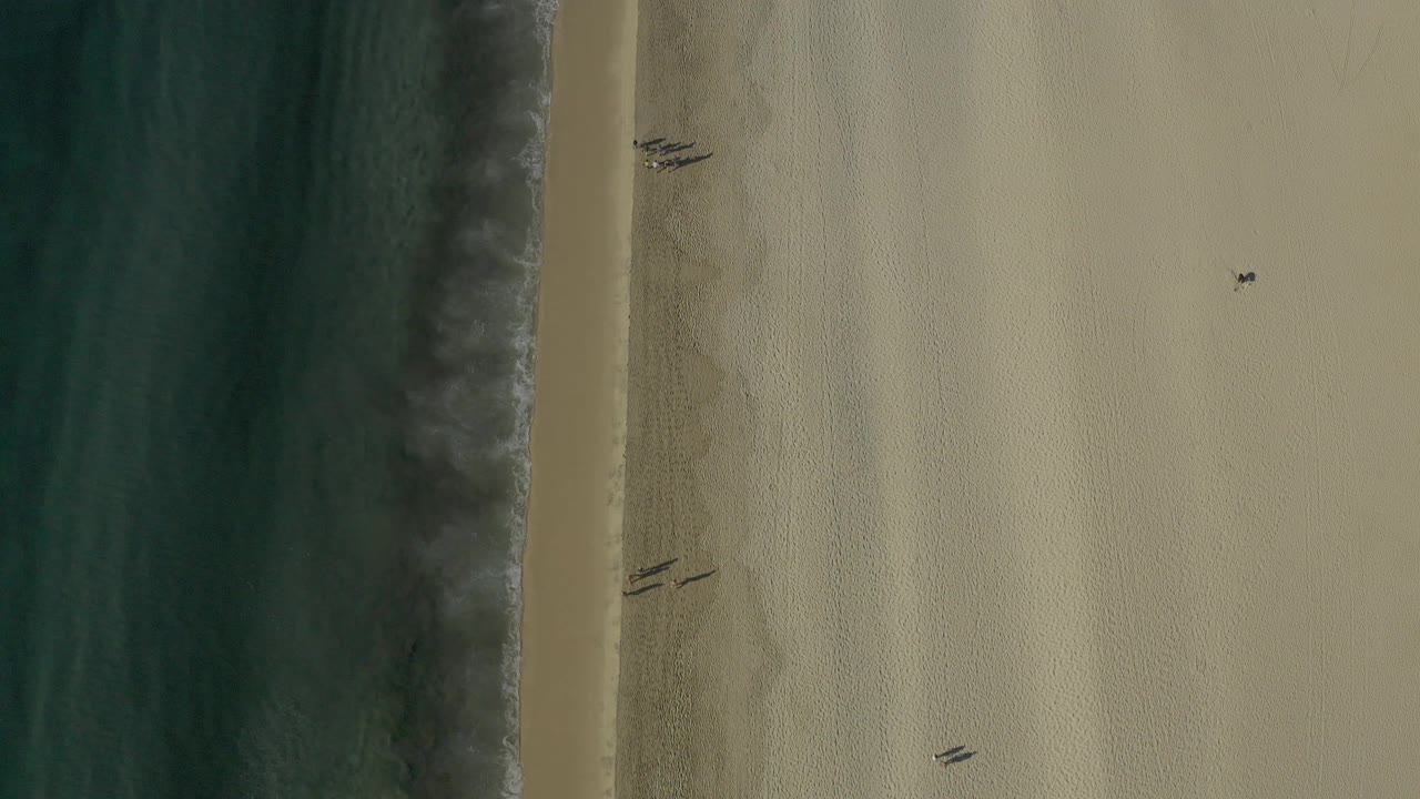 Overhead frame from Baja California Sur with dark water on the left, soft wave foam along the beach, small sand markings, and no visible people.