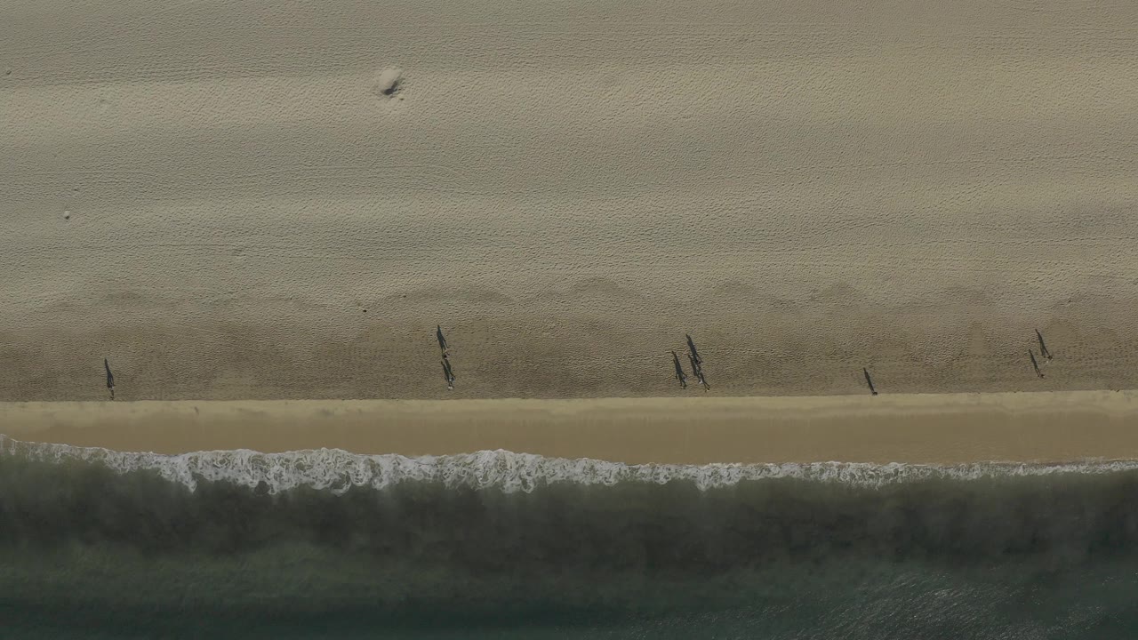 Overhead view of a Baja California Sur beach at 0:00, with dark green surf below the sand and small people walking near the waterline.