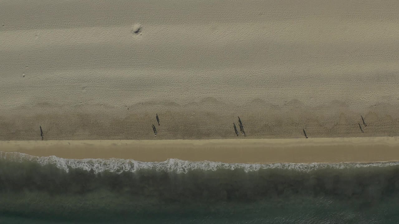 Top-down Baja California Sur shoreline at 0:02, with a darker water band along the bottom and several tiny figures moving across the open sand.
