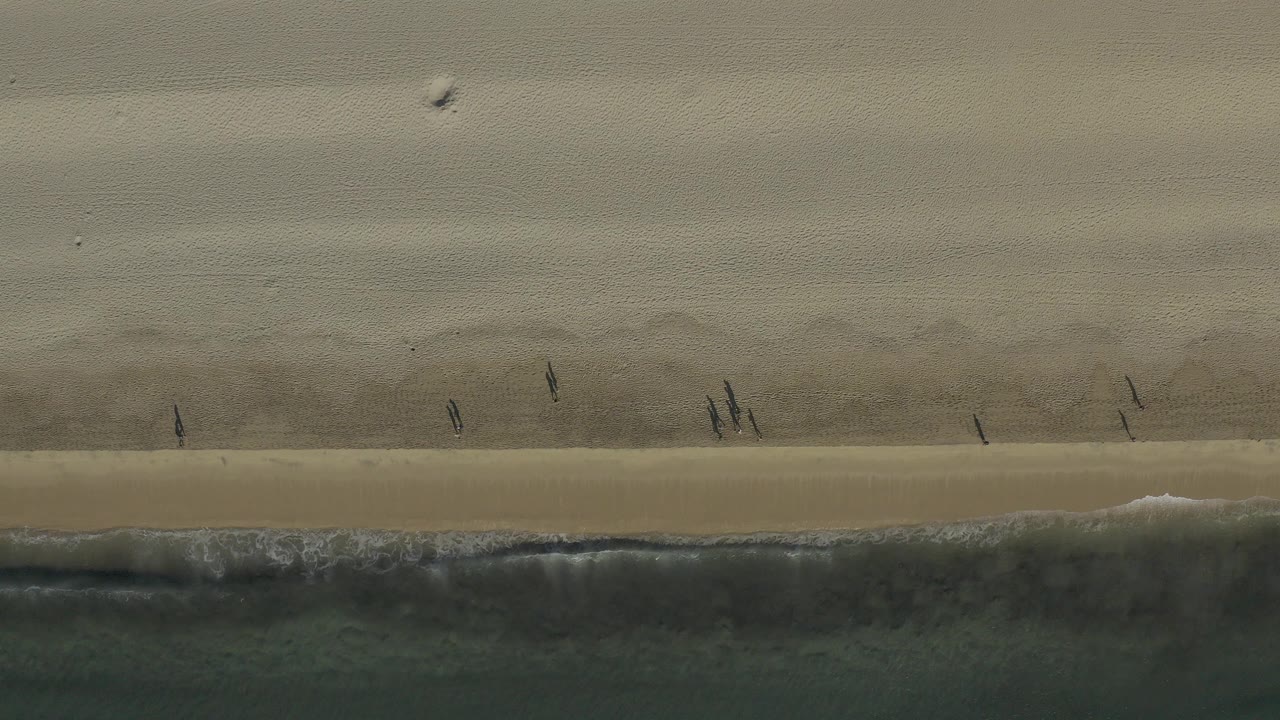 Overhead view in Baja California Sur, Mexico, showing pale sand, a narrow surf line, dark water, and several small people walking near the shoreline.