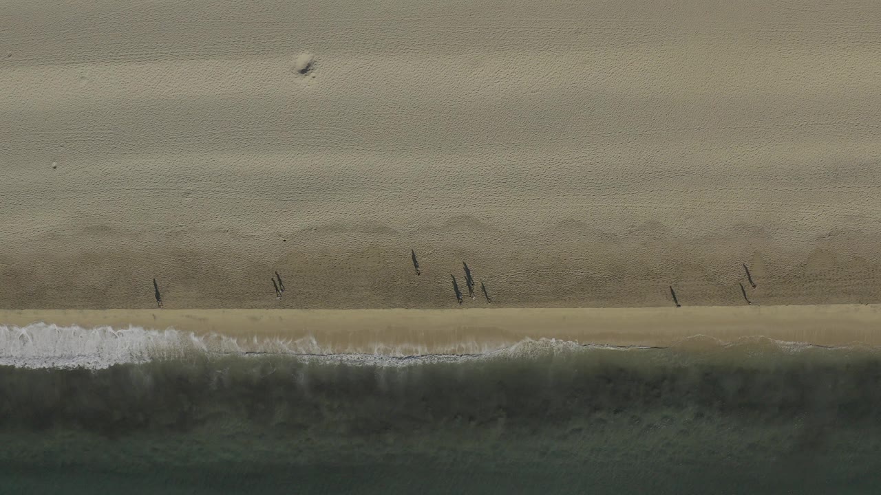 Overhead view of a Baja California Sur beach at 0:00, with tan sand above a dark green surf band and several small people near the shoreline.
