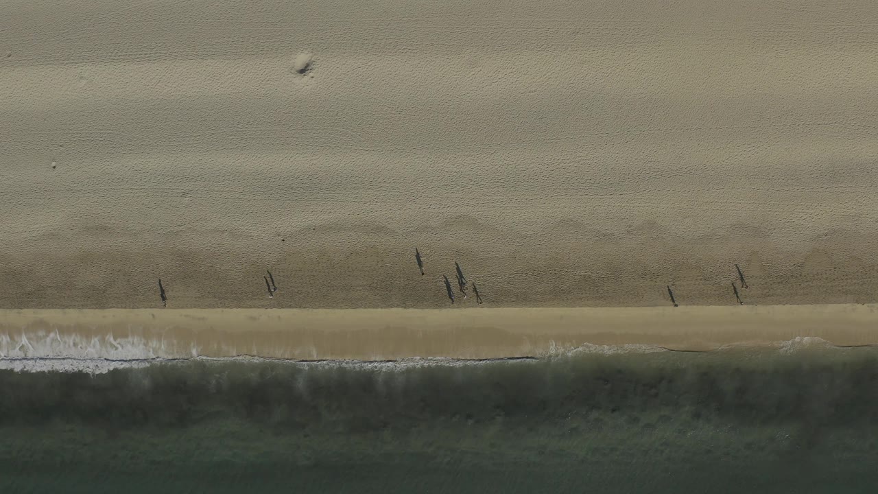 A top-down frame from Baja California Sur at 0:02 shows a narrow white surf edge, darker water below, and small figures standing or walking near the waterline.
