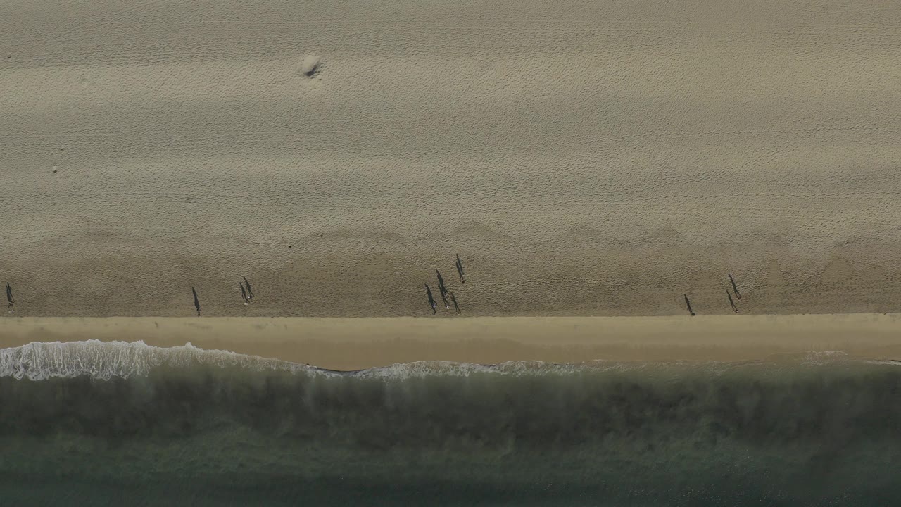 Overhead view of a Baja California Sur beach at the start of the clip, with pale sand above, green-gray surf below, and small people walking near the wet shoreline.