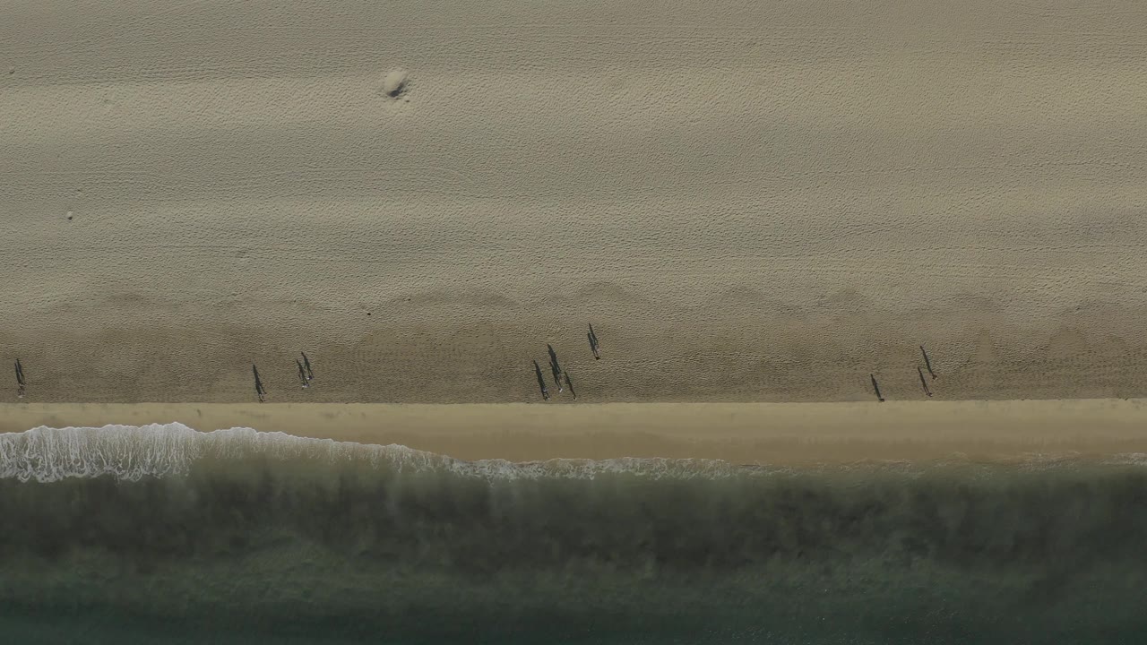 Top-down Baja California Sur beach view with small grouped walkers near the shoreline and a darker band of ocean pushing a thin foam edge onto the sand.
