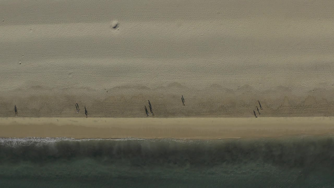 Overhead view of a Baja California Sur beach with pale sand, a dark wet shoreline band, green surf, and several small people walking near the waterline.