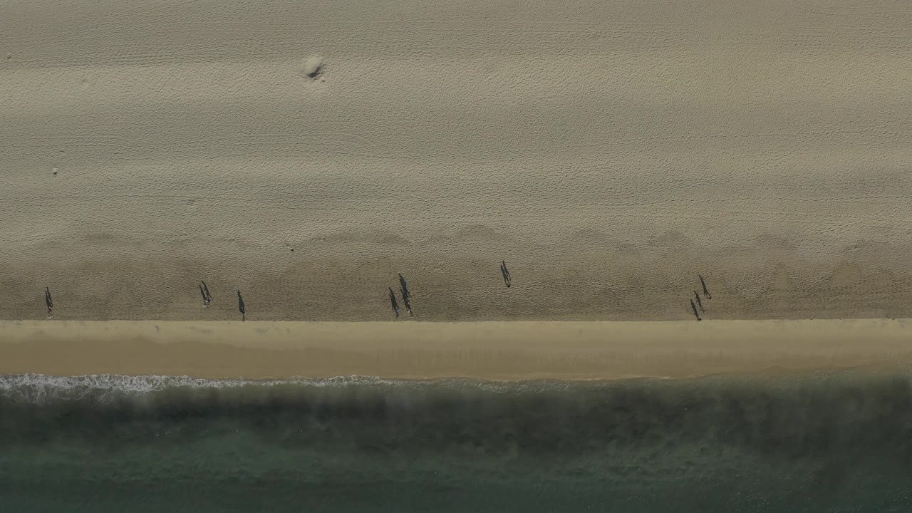 Aerial view over Baja California Sur sand and water, where small dark figures walk along the beach above a broad green-blue surf band.