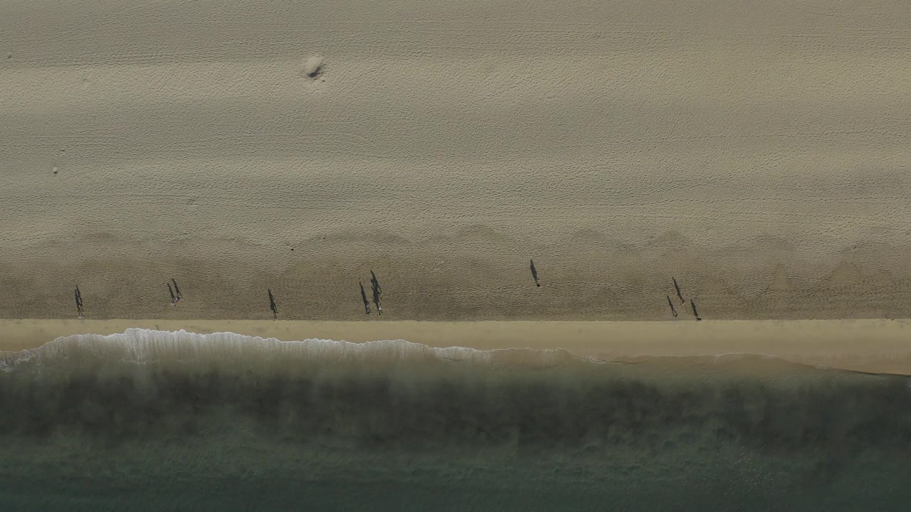 Top-down view of a Baja California Sur beach with pale sand, dark shoreline water, white surf, and several small walkers near the waterline.