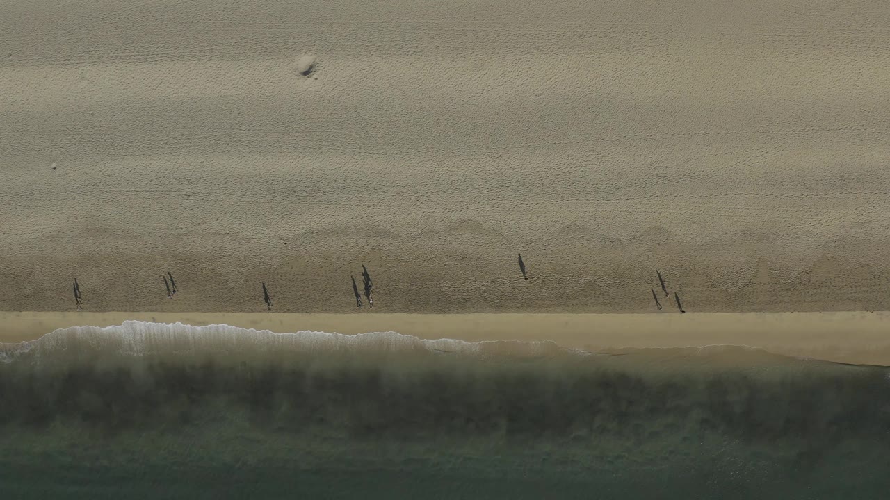 Overhead Baja California Sur shoreline frame with small figures in dark clothing spread across the sand above a narrow band of surf.