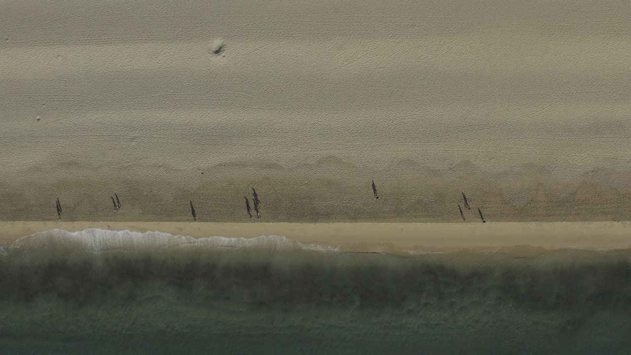 Straight-down beach view in Baja California Sur with the surf line crossing the lower frame and small people visible on the sand above it.