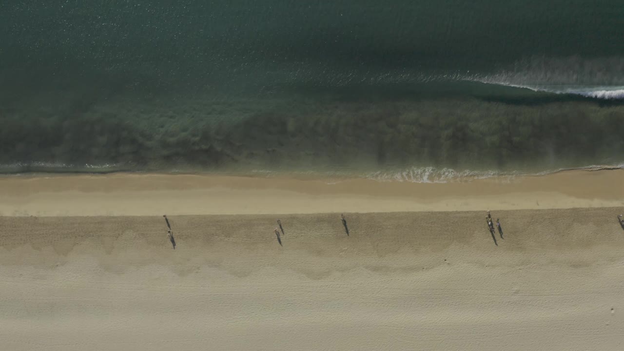 Overhead aerial frame in Baja California Sur, Mexico showing dark green ocean above a narrow surf line and several tiny people walking on the pale beach.