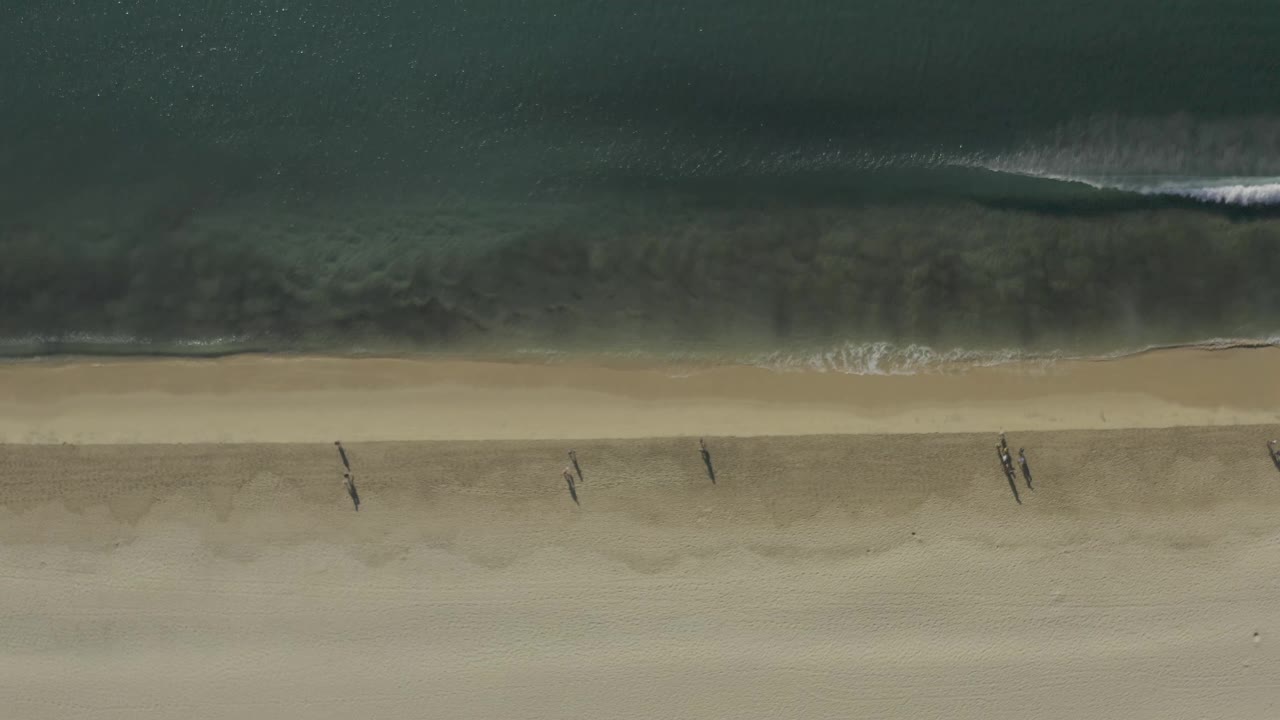 Aerial shoreline with small waves and walkers spaced across the sand.