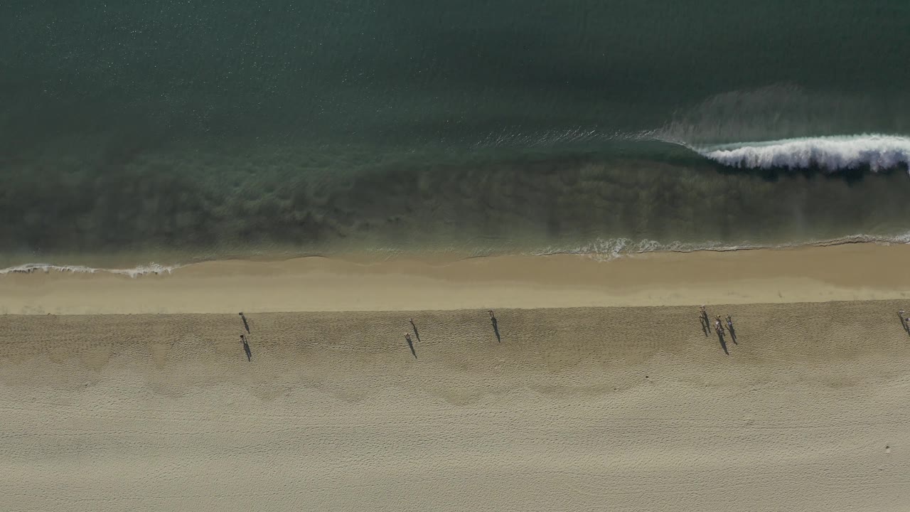 Top-down Baja California Sur shoreline frame where a brighter white wave starts breaking from the right while tiny walkers remain visible on the beach.