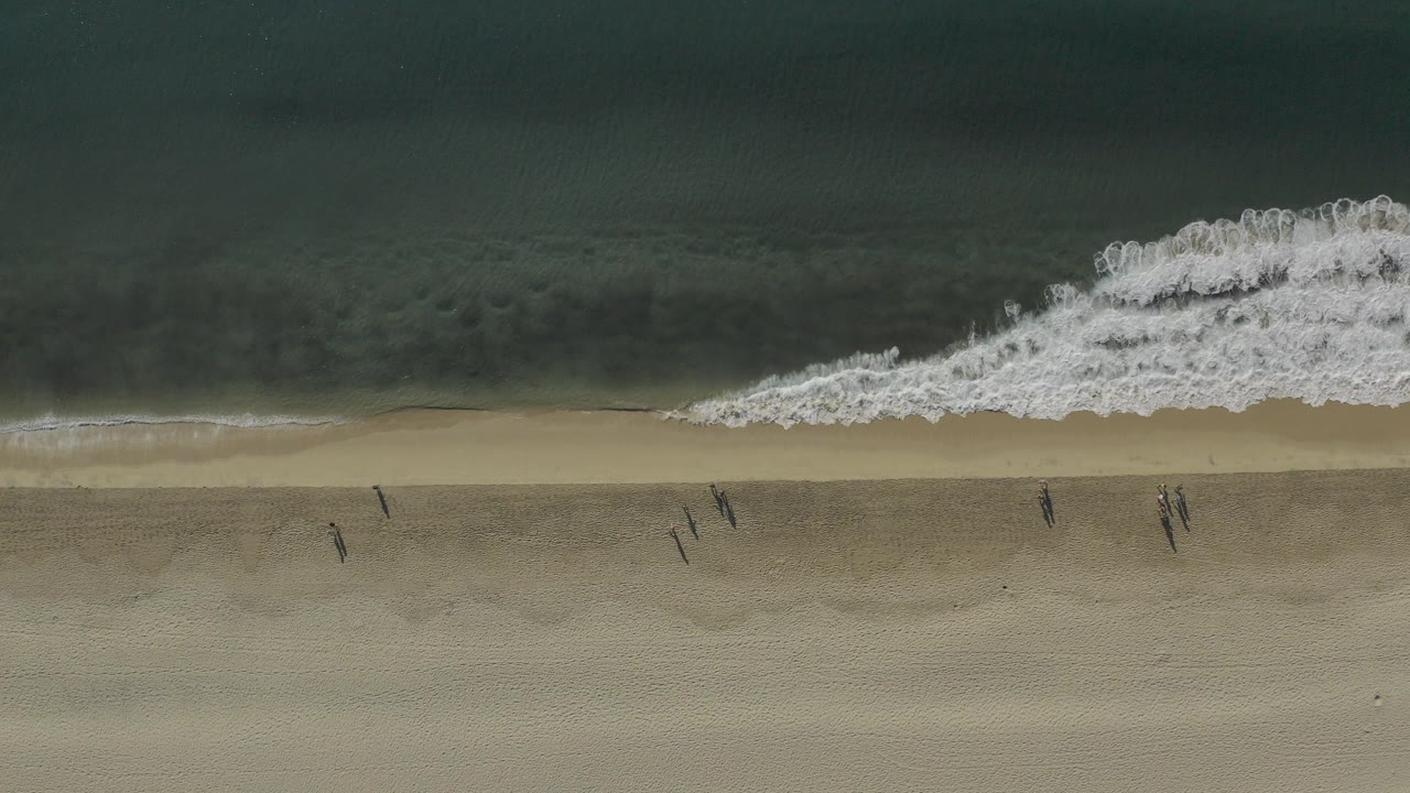 Overhead view in Baja California Sur, Mexico of a white wave beginning to break across dark green water and tan sand, with no clearly visible people.