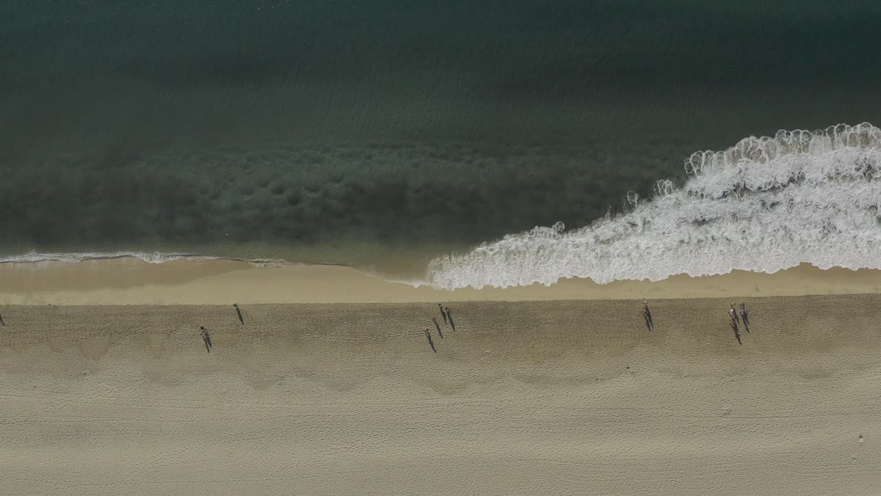 Overhead frame from Baja California Sur, Mexico showing a large white wave curling near the top of the shoreline over muted green water and tan beach, with no clearly visible people.