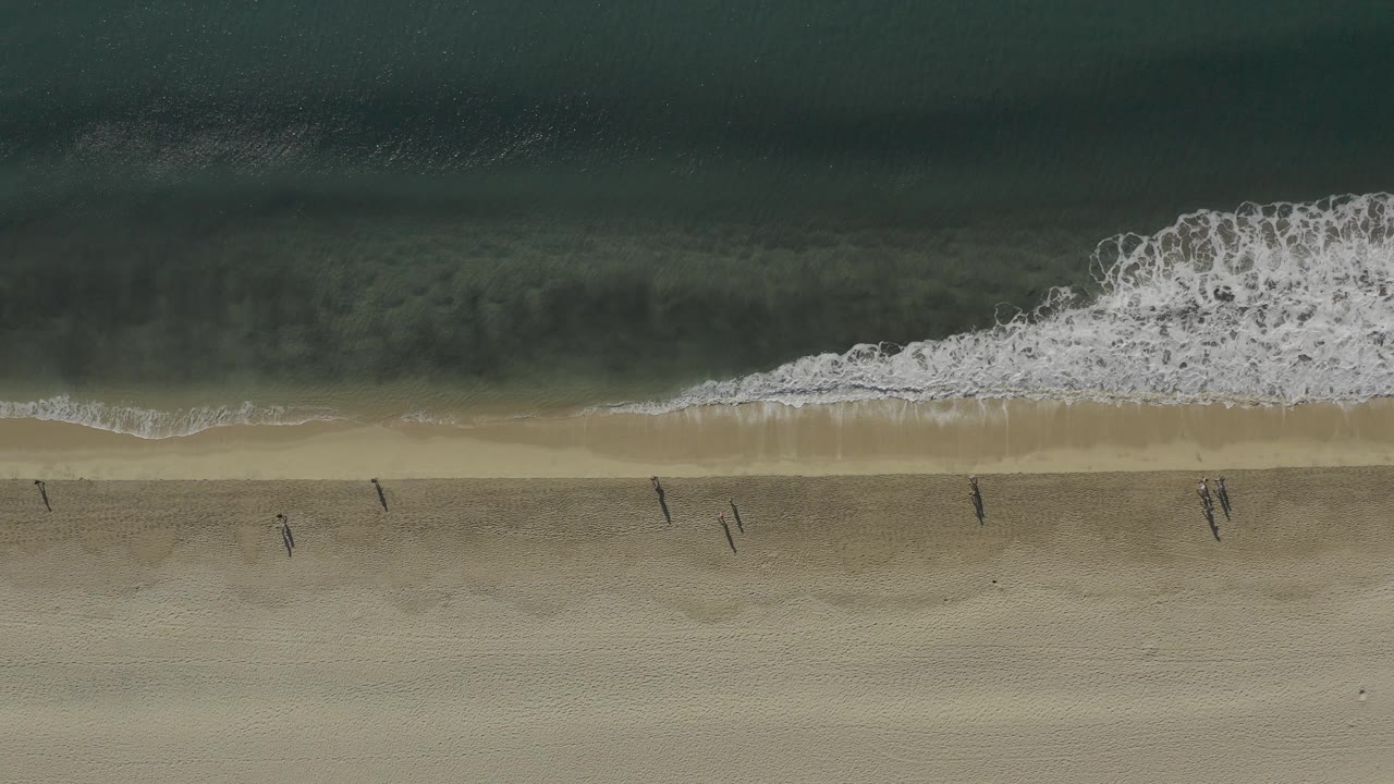 Top-down aerial frame in Baja California Sur, Mexico showing dark water, a white surf break, pale sand, and several small people walking near the shoreline.