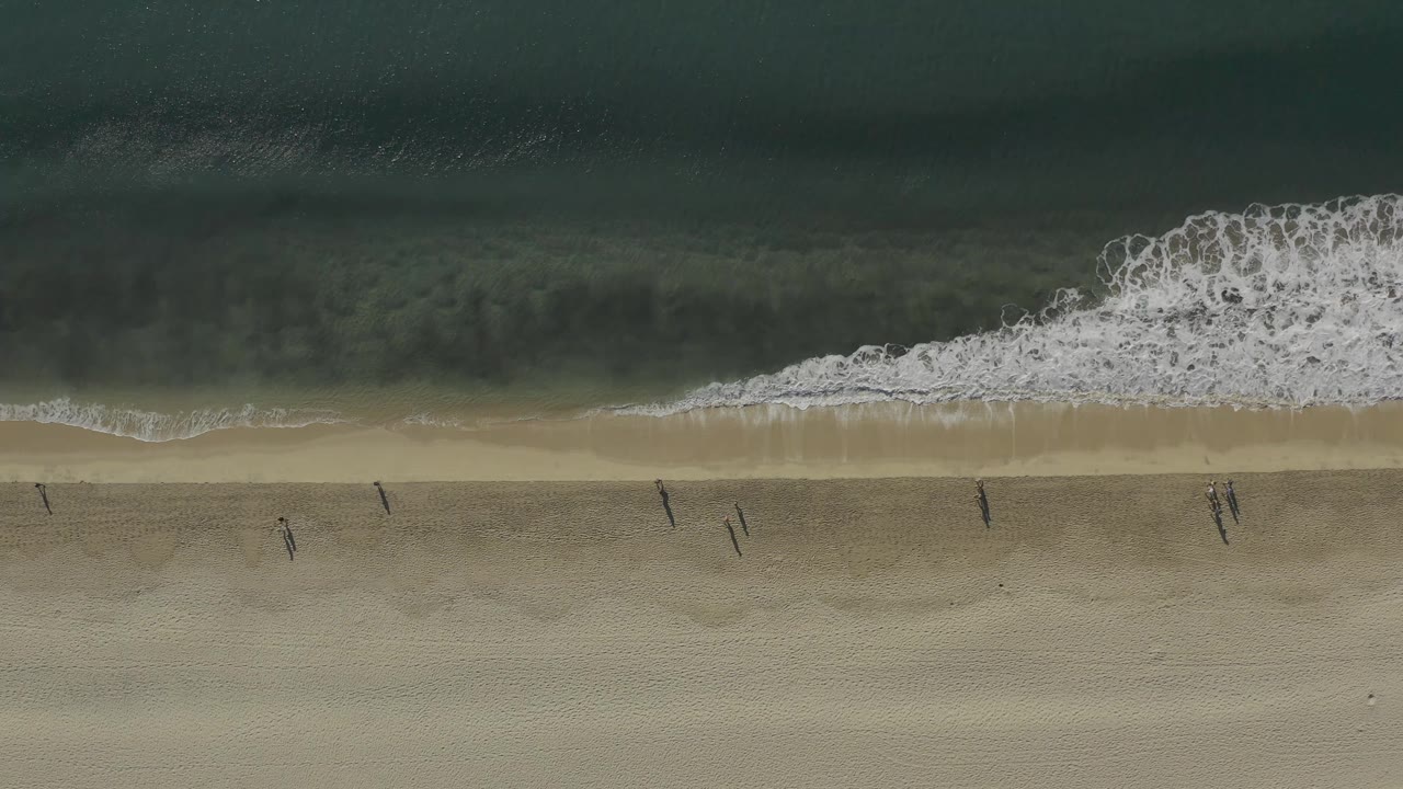 White surf curls onto sand as small figures walk along the beach.