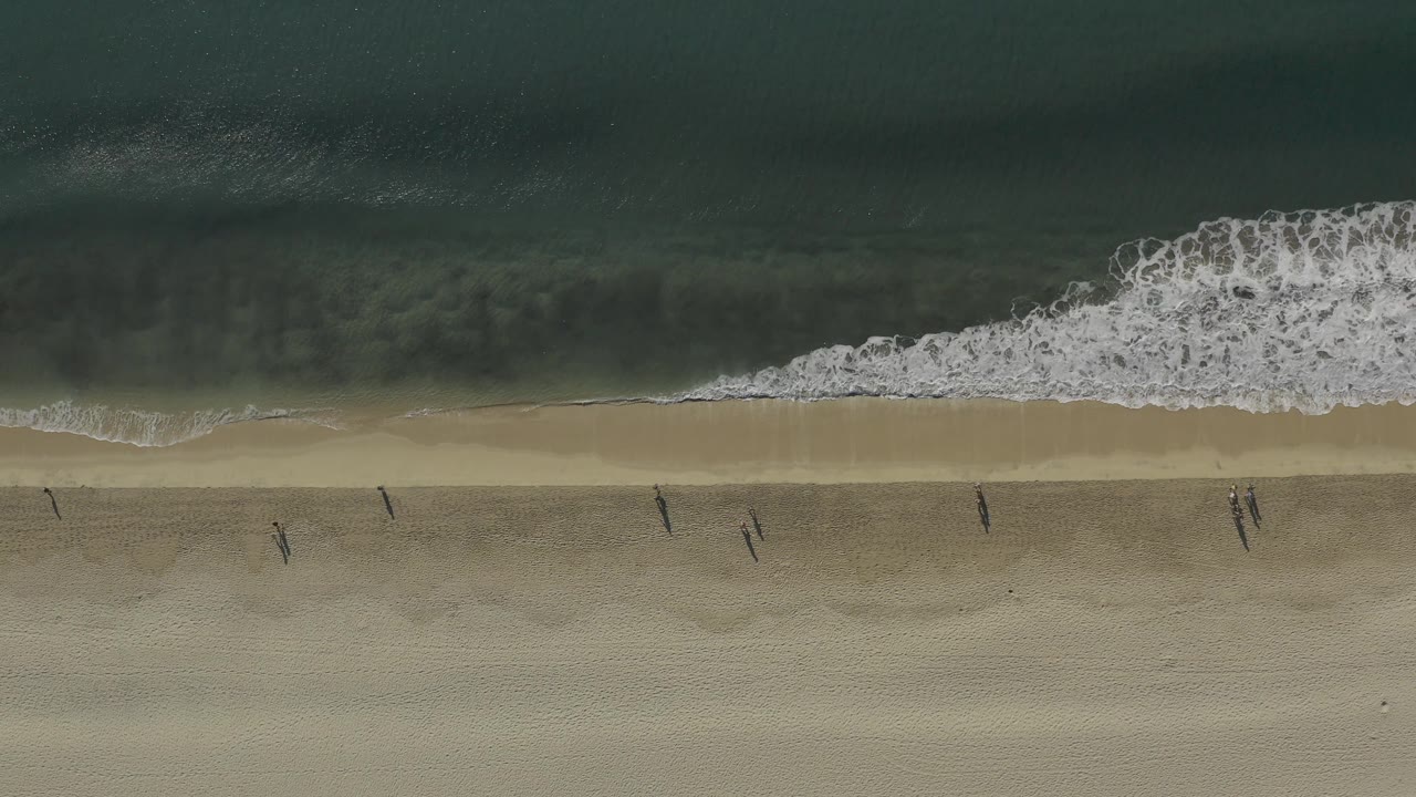 Overhead Baja California Sur beach view with a broken white wave near the top of frame and small people walking across the open sand below.