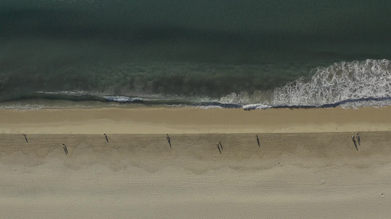 Overhead view in Baja California Sur, Mexico, showing green ocean, a narrow white surf line, pale sand, and small people spaced along the beach.