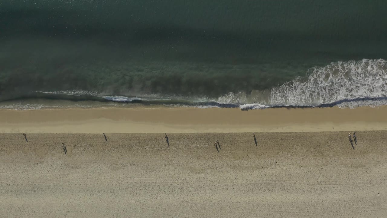 Aerial frame over Baja California Sur, Mexico, with breaking wave foam near the center and several tiny walkers casting shadows on the sand.