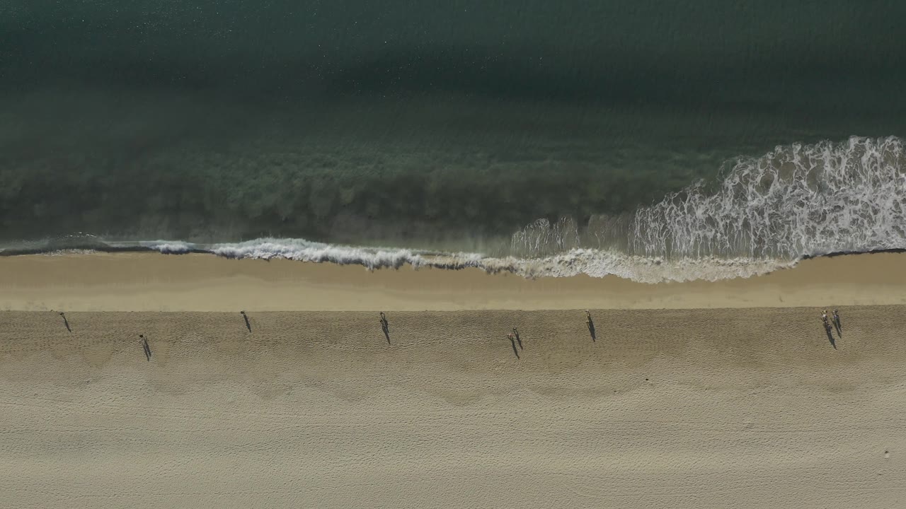 Top-down view in Baja California Sur, Mexico, where a larger patch of white foam breaks at frame right above a wide strip of sand with distant people.