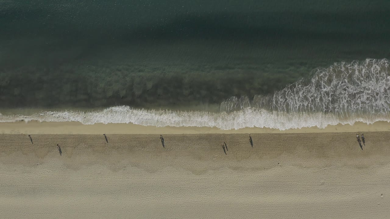Drone view of surf washing onto sand with small people below.