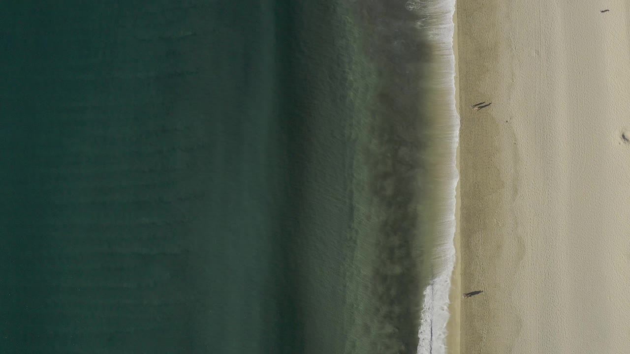 Overhead view in Baja California Sur, Mexico, with dark green ocean at left, pale sand at right, a thin foam line, and two small people walking near the shoreline.