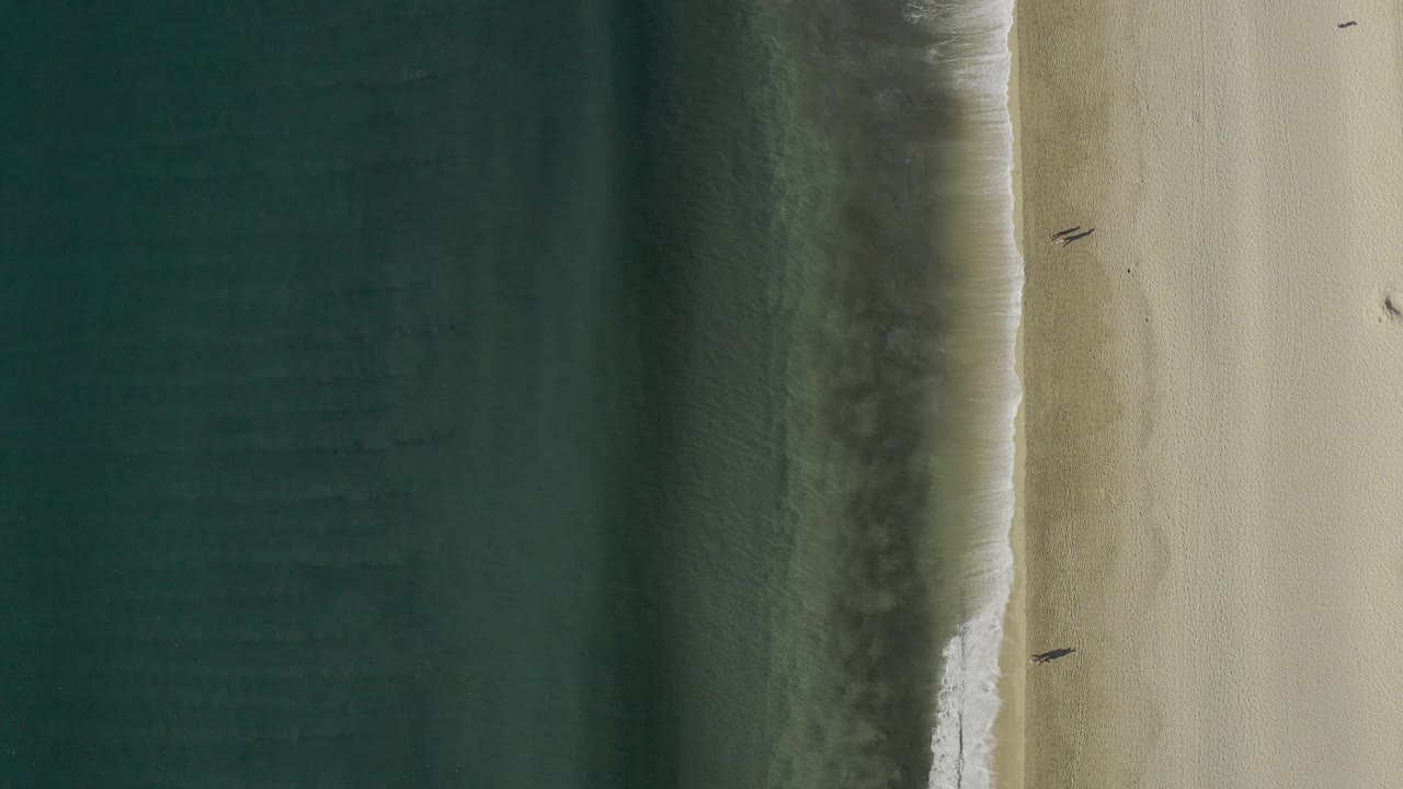 Aerial Baja California Sur shoreline frame showing the surf line slightly lower in the composition, two distant people on the beach, and broad empty sand around them.