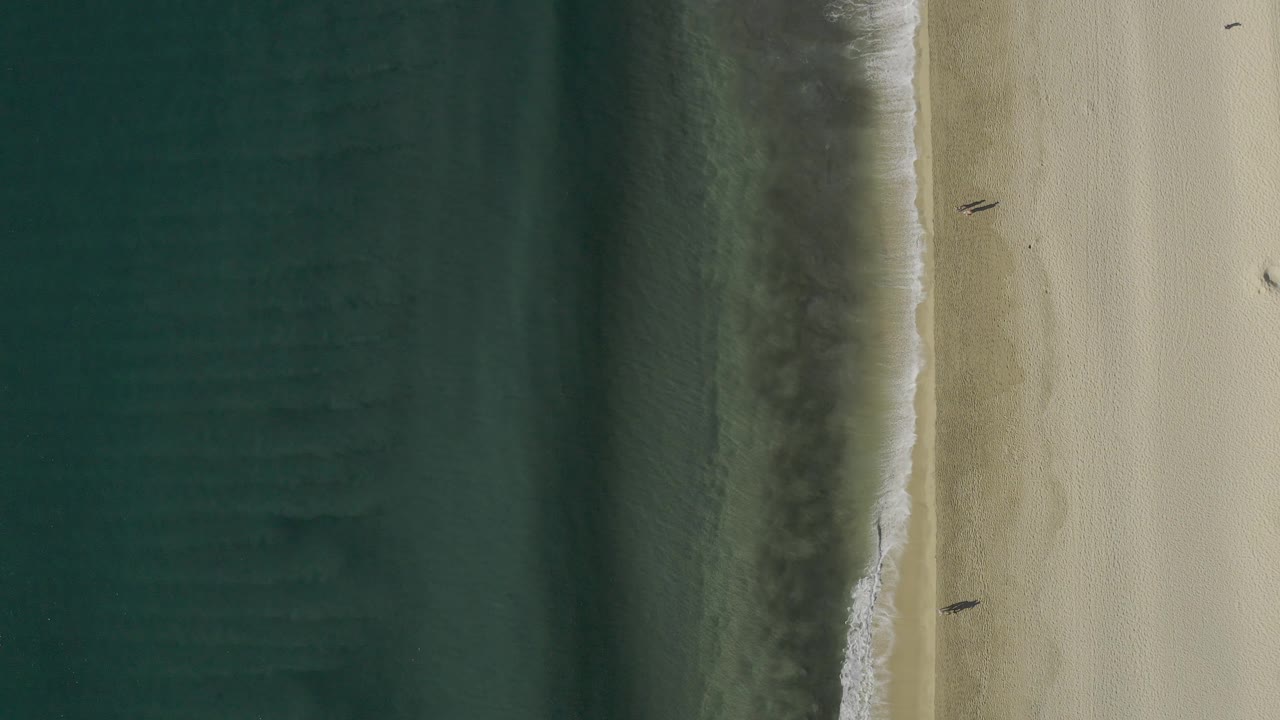 Overhead frame from Baja California Sur, Mexico, with textured green water, a narrow white wave edge, pale beach, and two tiny walking figures separated along the sand.