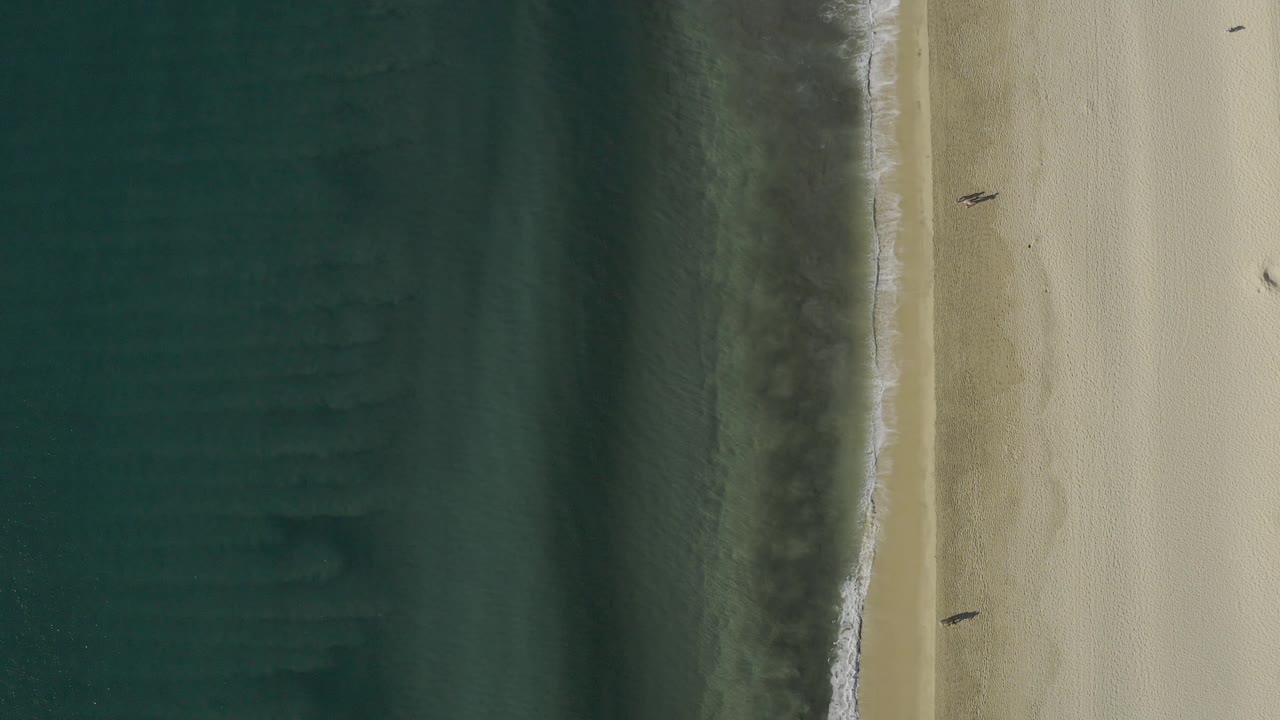 Aerial surf edge and sand with two tiny beach walkers visible.