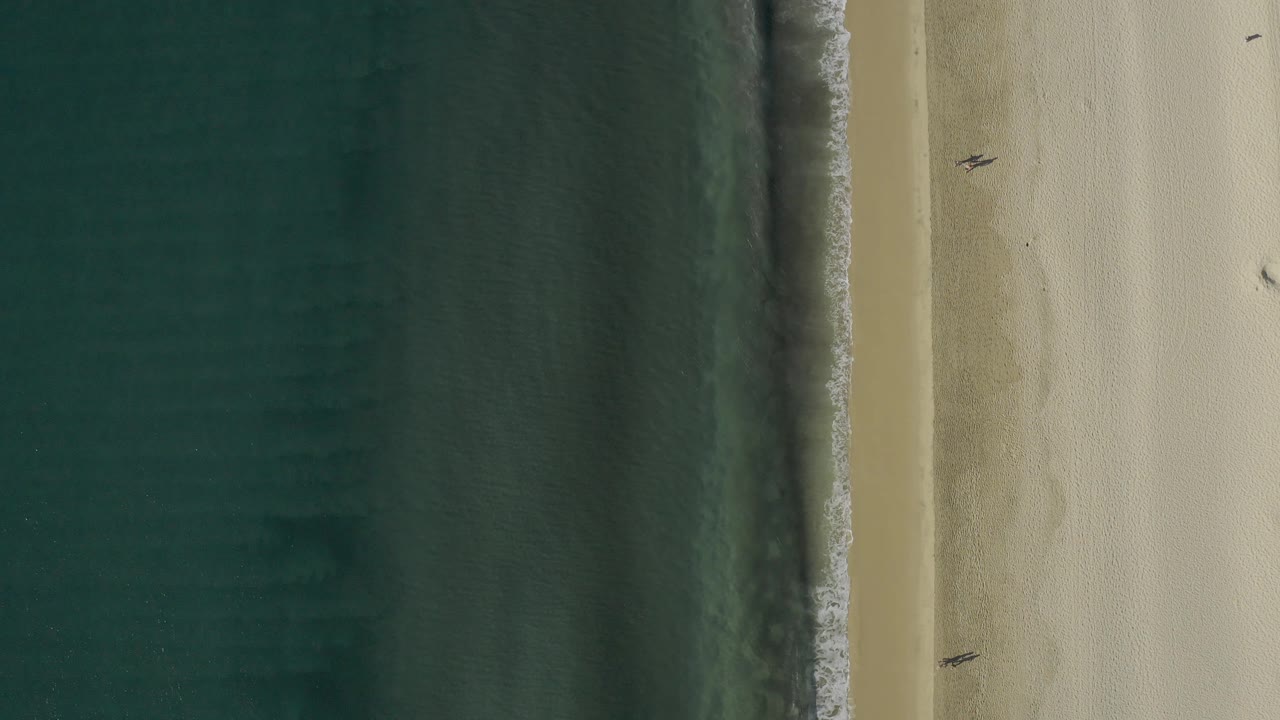 High drone view in Baja California Sur, Mexico, with darker water texture on the left, a bright foam edge, broad sand on the right, and tiny people visible near the beach side.