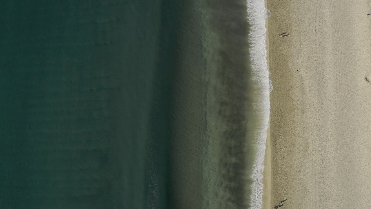 Straight-down drone frame over Baja California Sur, Mexico, where a darker wet band separates teal water from pale sand, with no visible people.