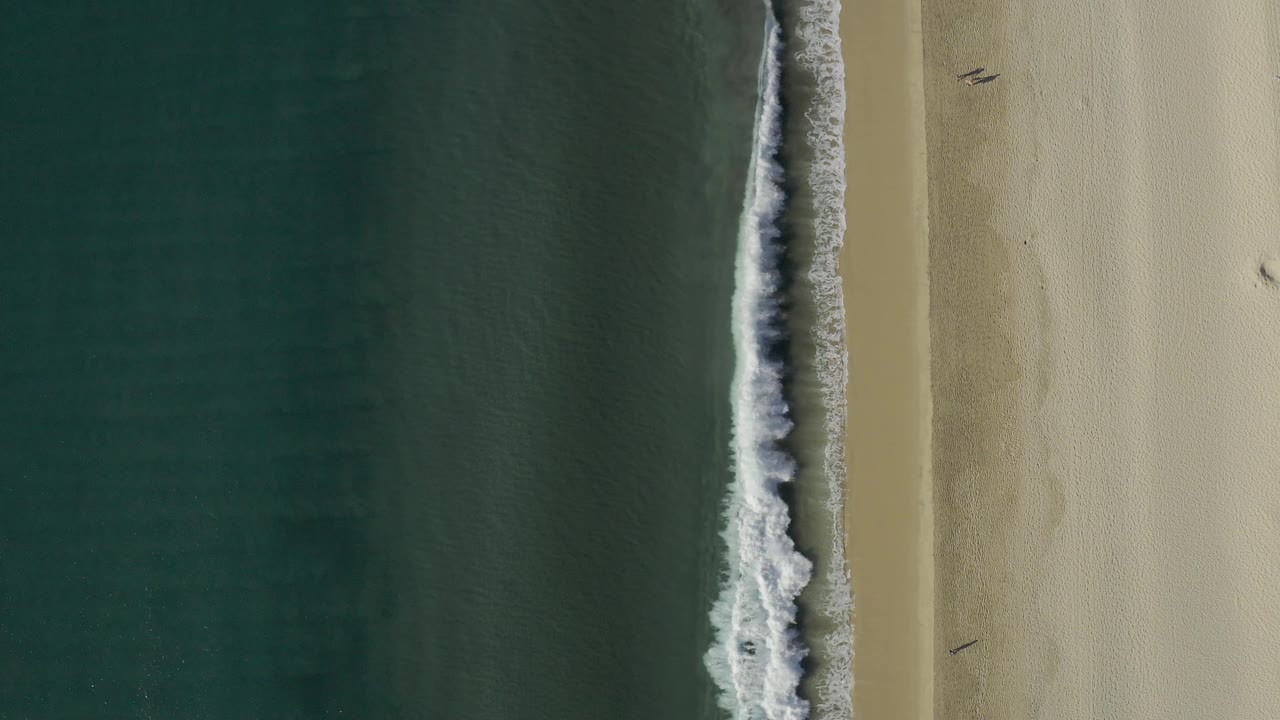 Aerial frame over Baja California Sur coast where white foam spreads wider along the wet shoreline between green water and pale sand, with no visible people.